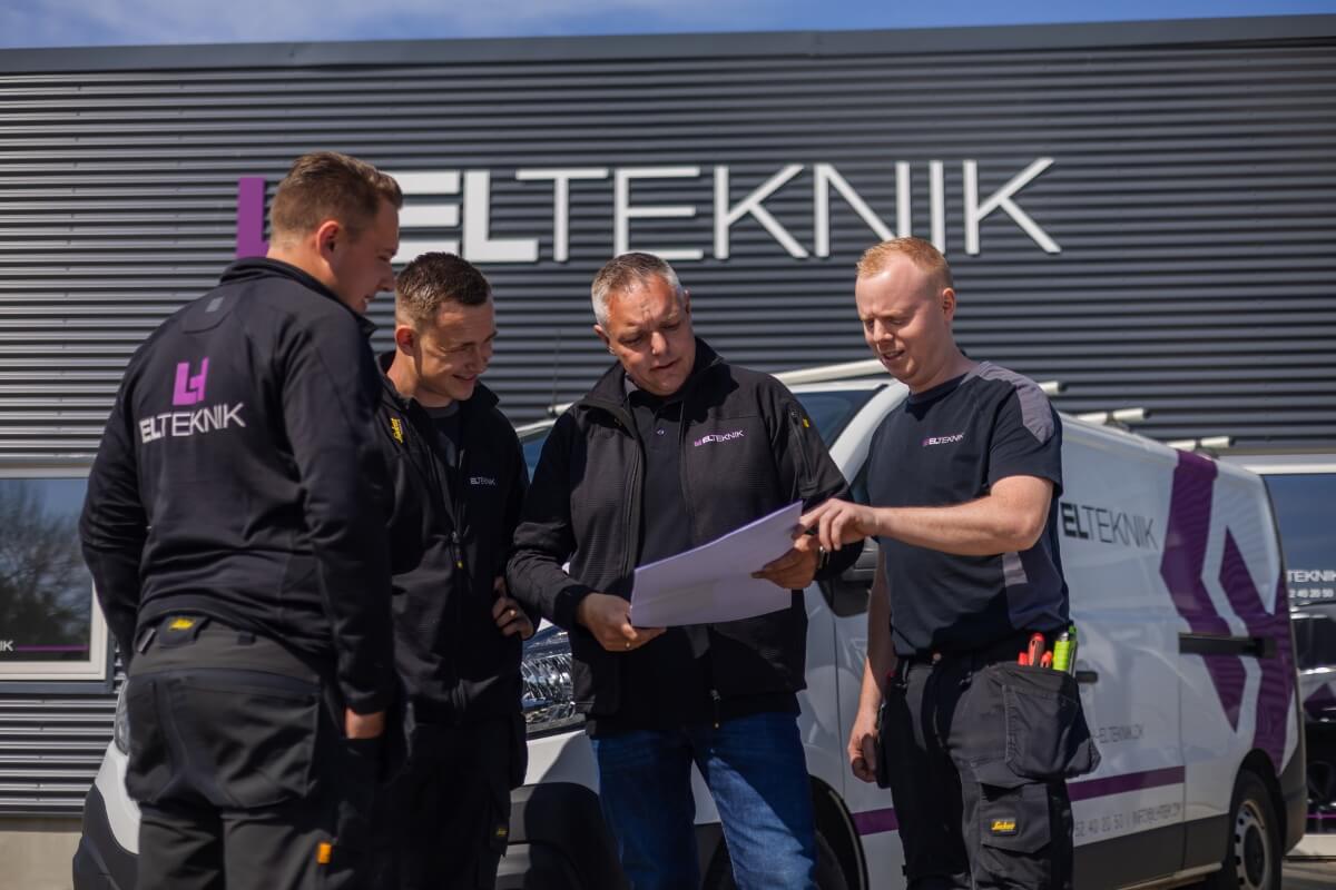 Fire elektrikere fra ELTEKNIK står foran en servicebil og gennemgår tegninger sammen foran virksomhedens bygning med logo i baggrunden. Situationen viser samarbejde og planlægning af en opgave inden udførelse.