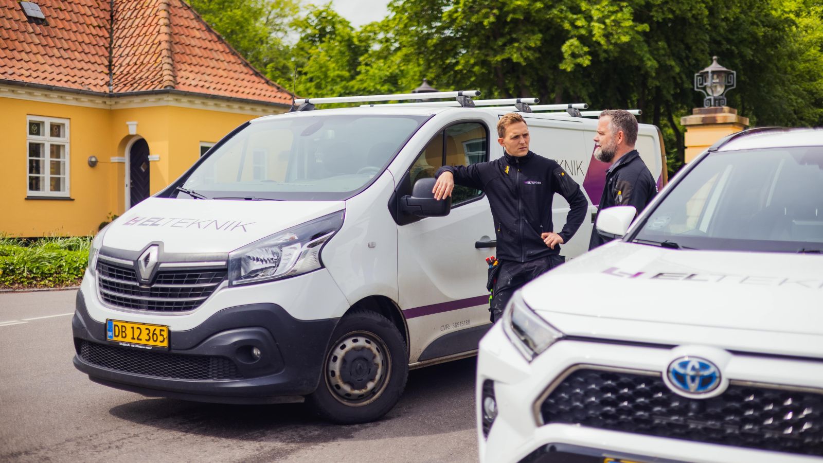 To elektrikere fra ELTEKNIK står ved deres firmabiler og taler sammen på en villavej foran et gult hus. De planlægger dagens opgaver med værktøj i bæltet og servicebiler klar til kørsel.