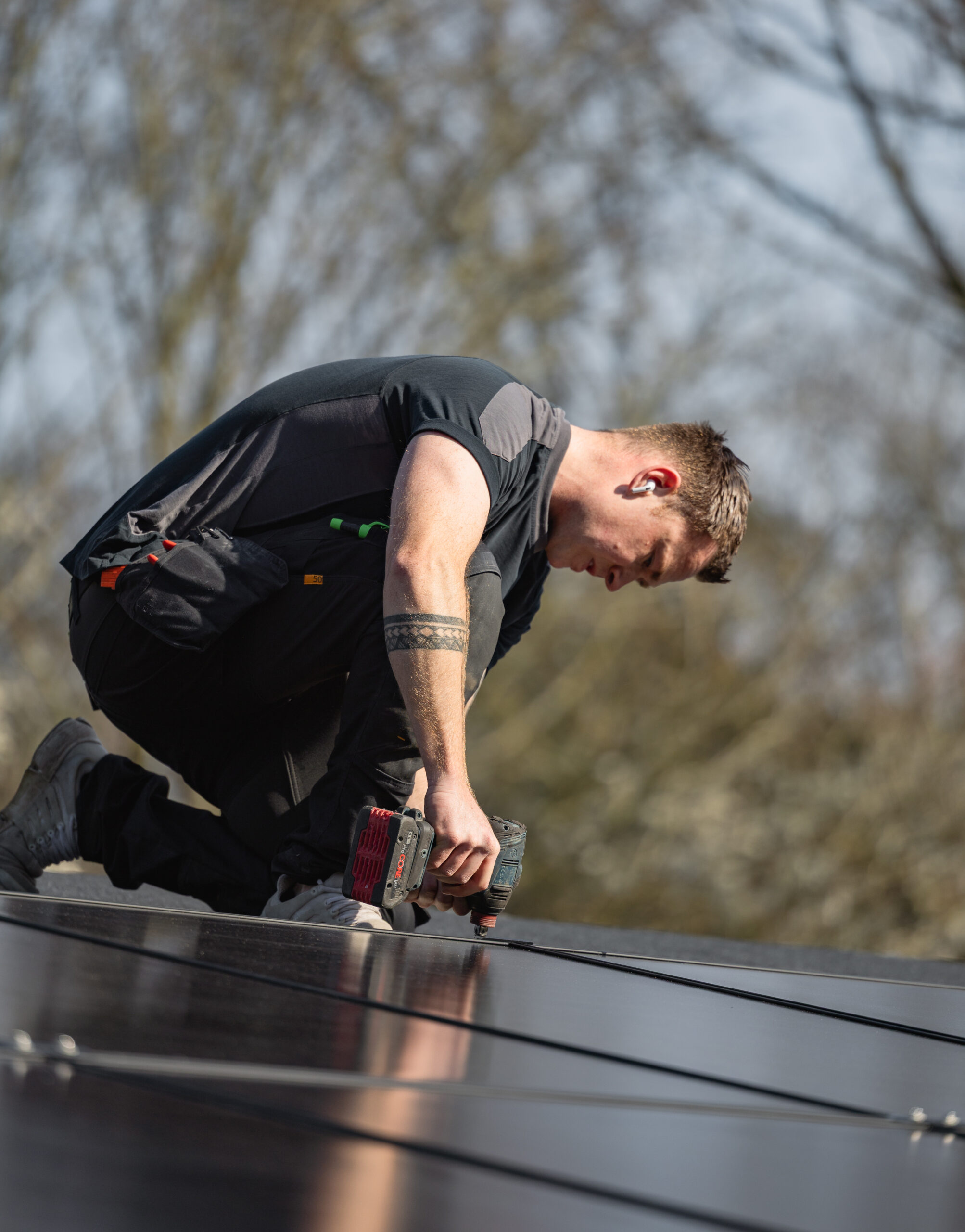 Elektriker knæler på tag og fastgør solcellepanel med boremaskine som en del af installation af solcelleanlæg. Arbejdet viser præcis montering af paneler der skal producere strøm fra solenergi.