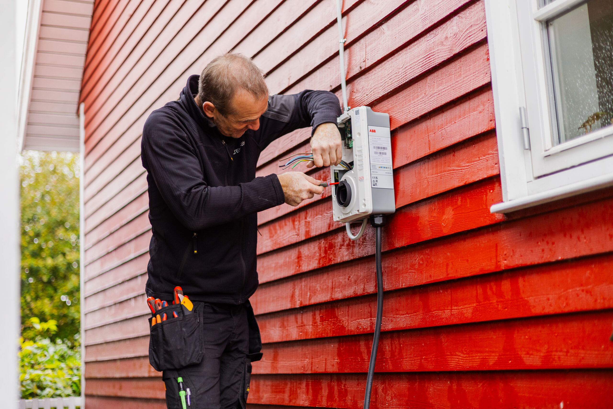 Elektriker fra LH Elteknik monterer og tilslutter ledninger i en udendørs elboks på en rød træfacade ved en bolig. Billedet viser præcisionsarbejde med installation af eludstyr.