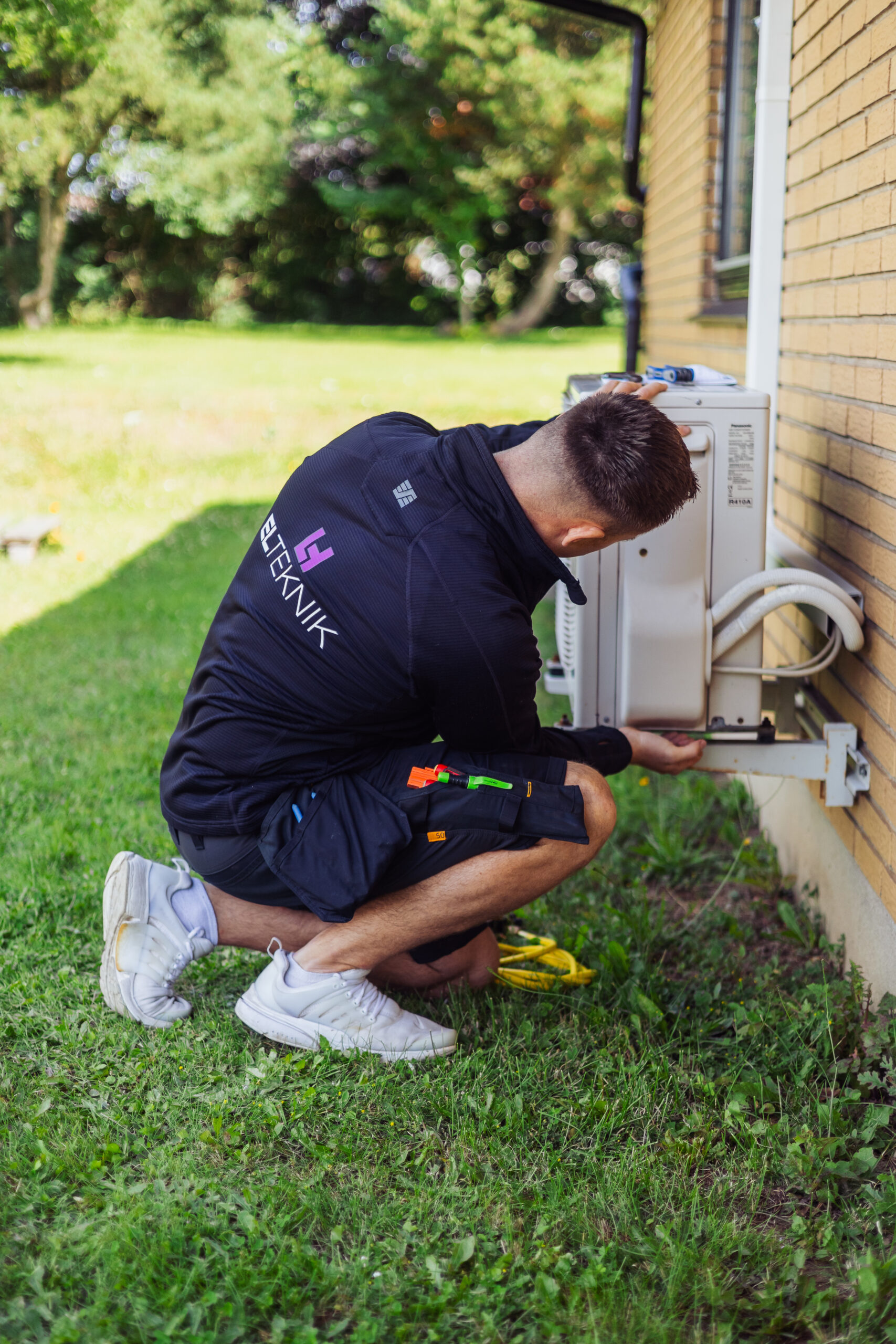 Elektriker fra LH Elteknik sidder ved husets facade og arbejder på en udendørs varmepumpe mens han justerer tilslutninger og kabler. Billedet viser service eller installation af varmepumpe i en have ved en bolig.