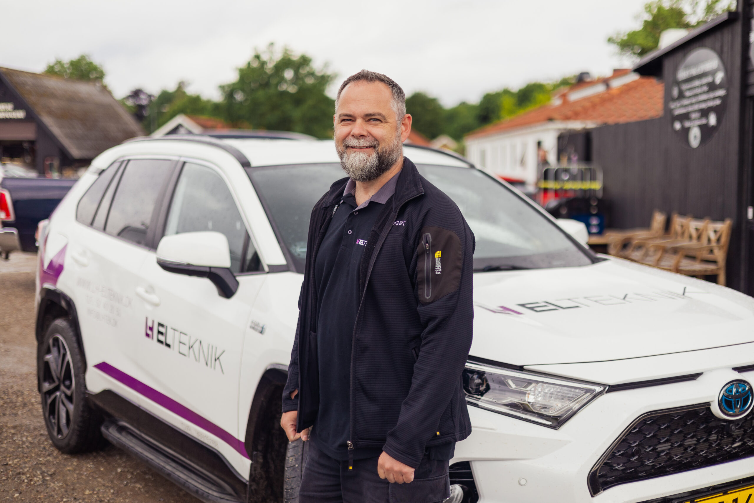 Smilende elektriker fra LH Elteknik står foran en hvid firmabil med logoet "ELTEKNIK" i et udendørs miljø med bygninger og træer i baggrunden. Hans arbejdstøj og bilen understreger hans rolle som elektriker på opgave.