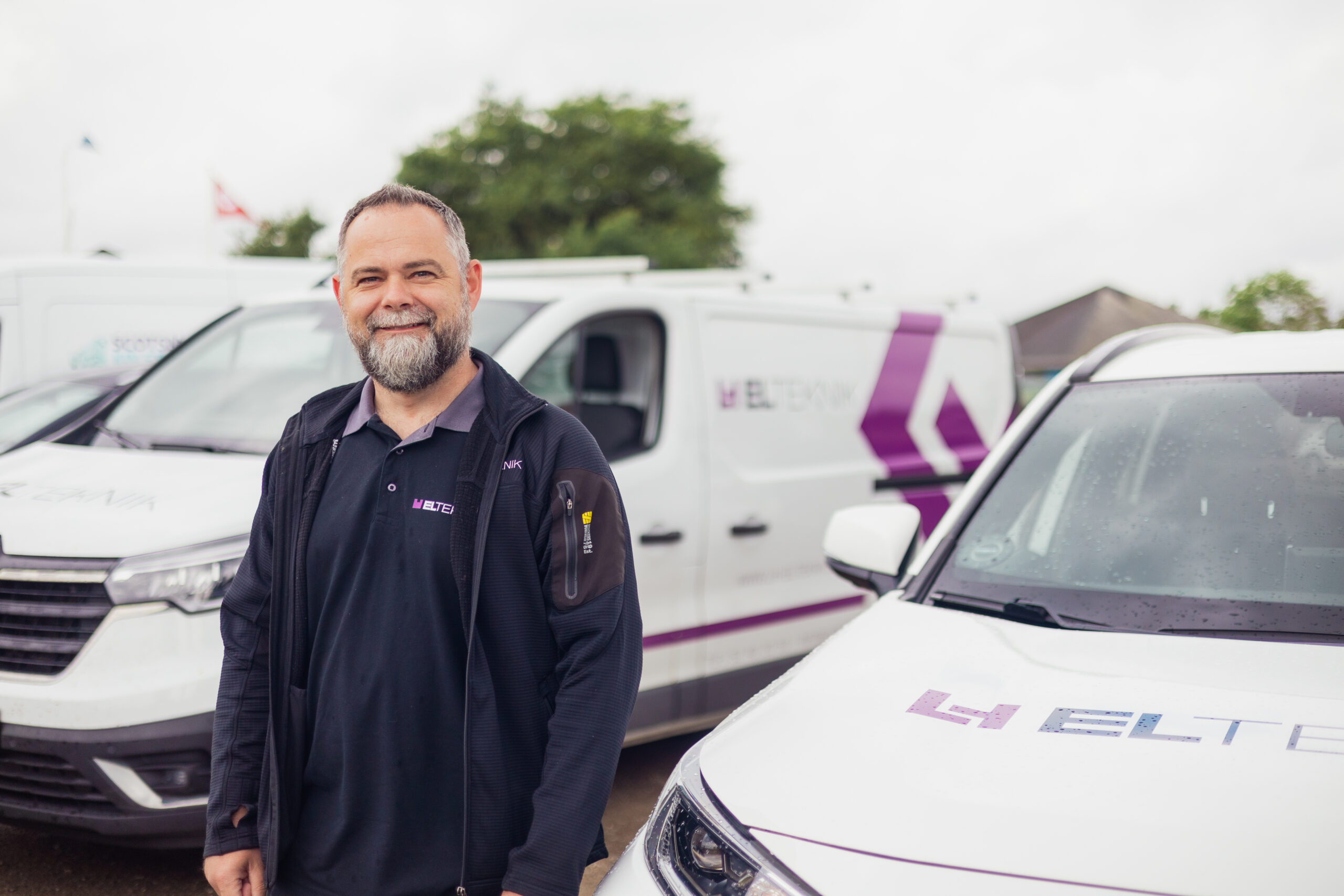 Elektriker fra LH Elteknik står foran firmabiler med logo og smiler mod kameraet på en overskyet dag. Billedet viser virksomhedens servicebiler og giver et professionelt indtryk af elektrikeren på arbejde.