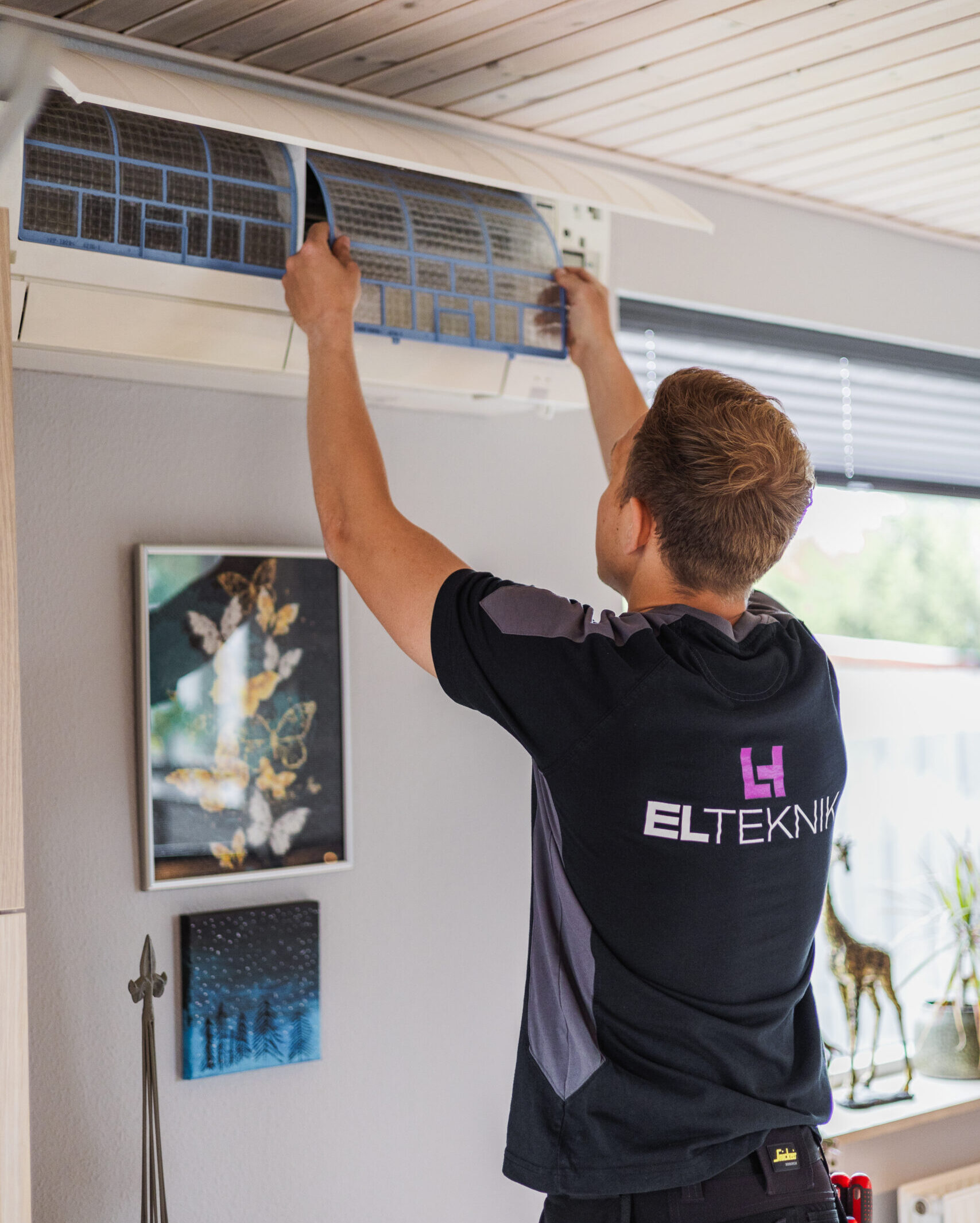 Elektriker fra LH Elteknik rengør eller udskifter filter i en vægmonteret varmepumpe indendørs. Arbejdet ved enheden viser service og vedligeholdelse som en del af opgaver for en elektriker.