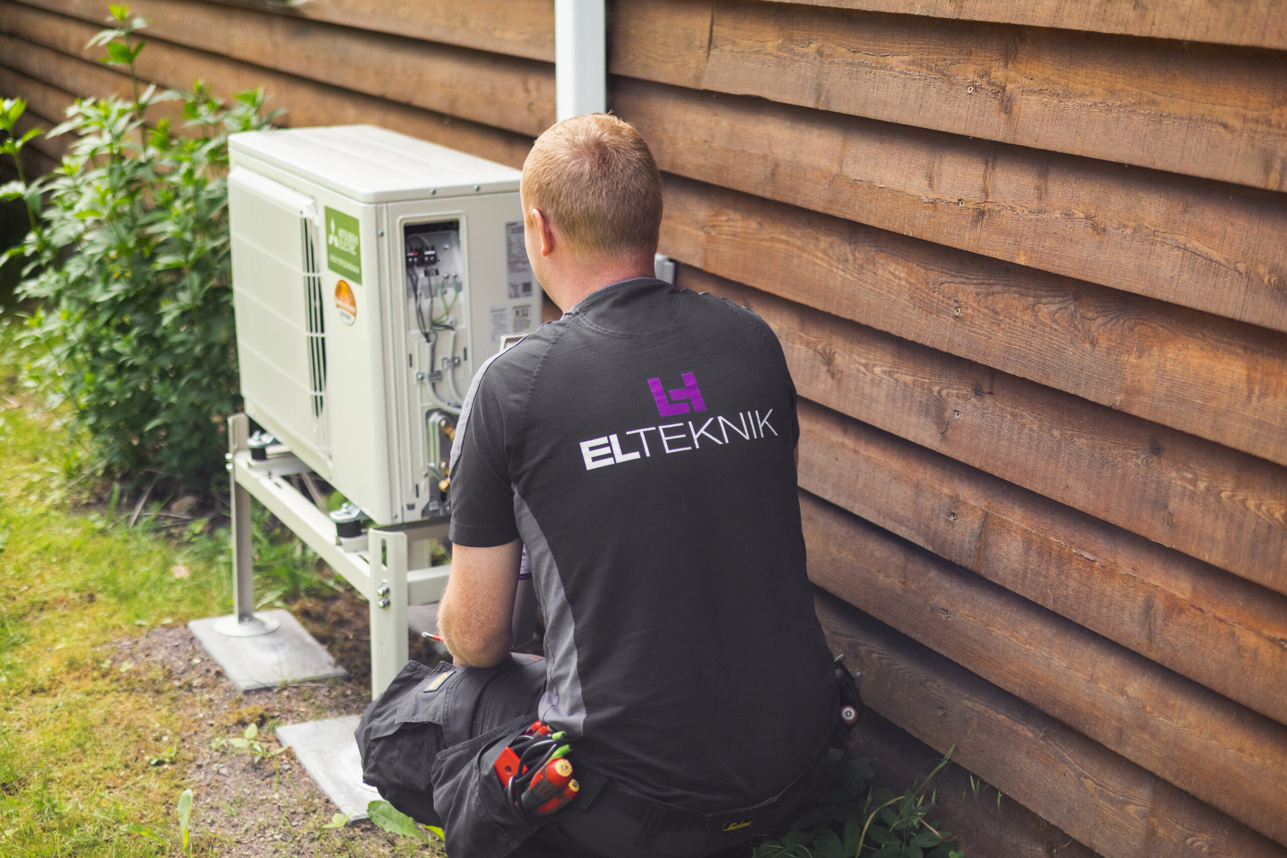 Elektriker fra LH Elteknik arbejder på en udendørs varmepumpe monteret ved en træfacade og justerer installationen. Billedet viser service og vedligeholdelse af varmepumpe ved bolig.
