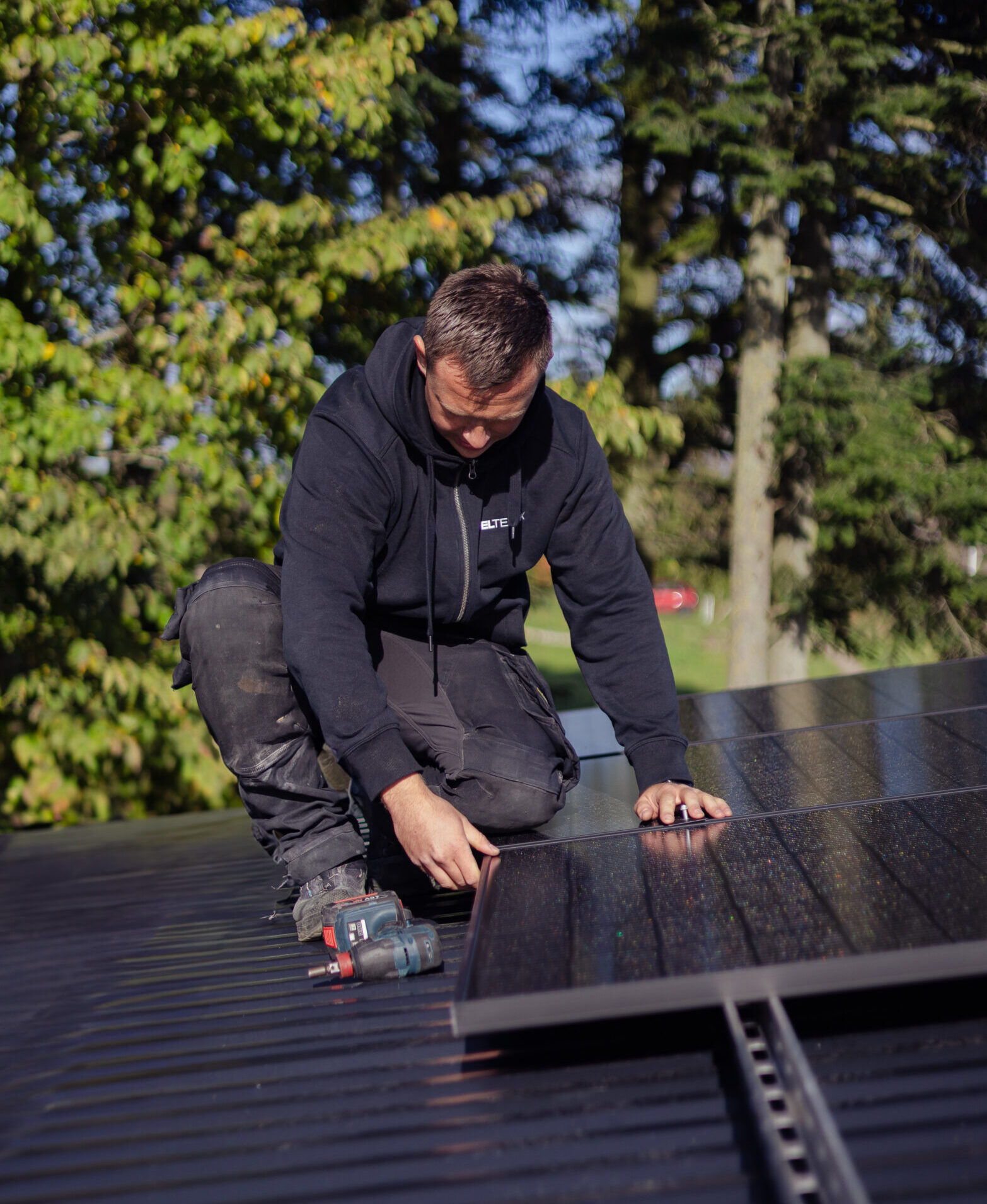 Elektriker fra LH Elteknik knæler på et tag og monterer solpanel med en boremaskine ved siden af sig. Installationen foregår udendørs i sollys med træer i baggrunden og viser opsætning af solcelleanlæg.