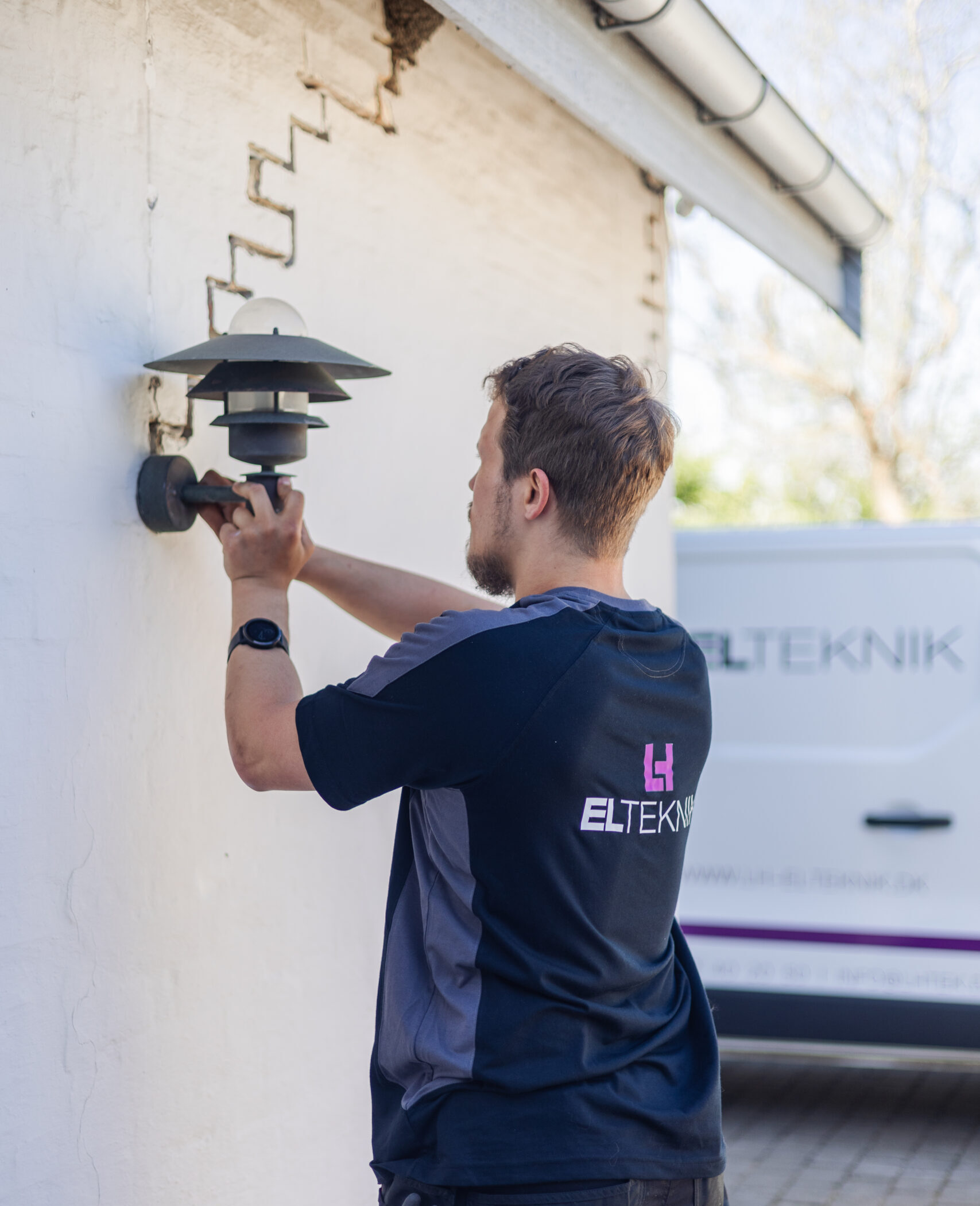 Elektriker fra LH Elteknik monterer en udendørs væglampe på en husfacade mens en servicebil holder parkeret i baggrunden. Arbejdet viser installation af belysning som en del af hans opgaver som elektriker.