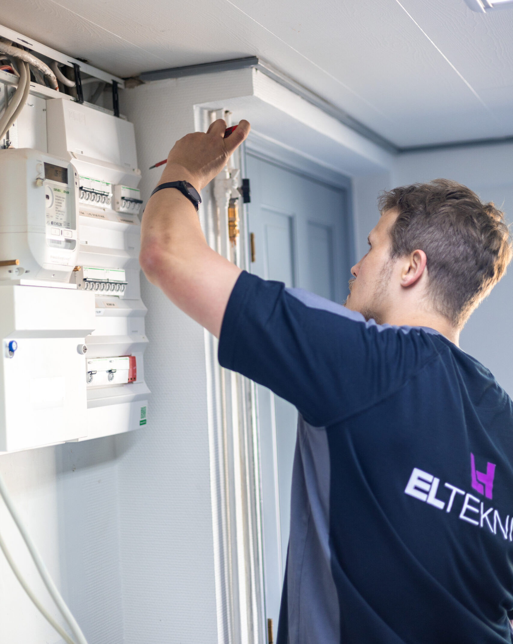 Elektriker fra LH Elteknik arbejder ved en eltavle indendørs og justerer installationer med en skruetrækker. De synlige sikringer og kabler viser præcisionsarbejde med elinstallation i et hjem.