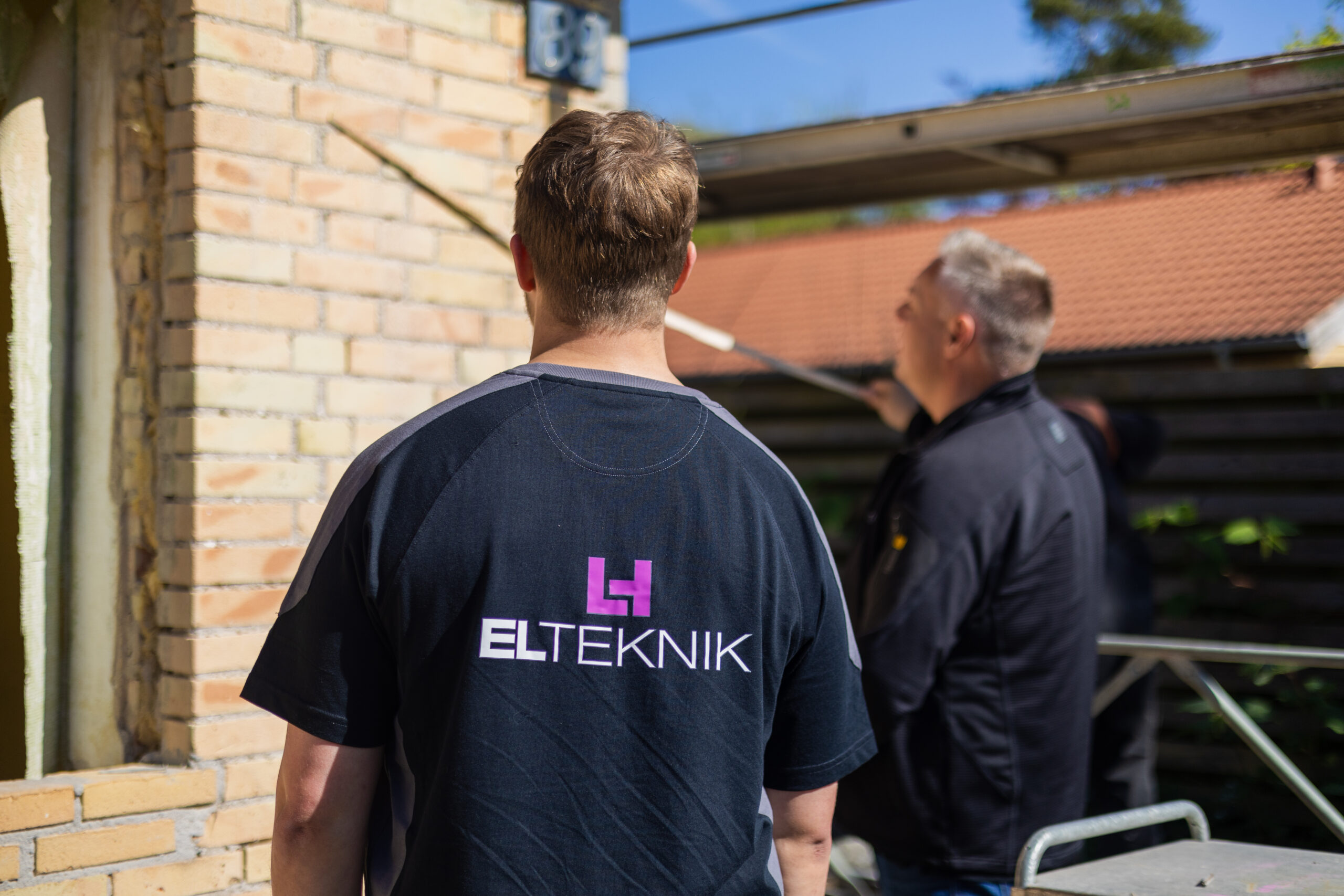 Elektriker fra LH Elteknik står med ryggen til mens en kollega arbejder med installation under et udhæng ved en murstensbygning. Logoet "ELTEKNIK" ses tydeligt på trøjen og viser dem i gang med en opgave som elektrikere.
