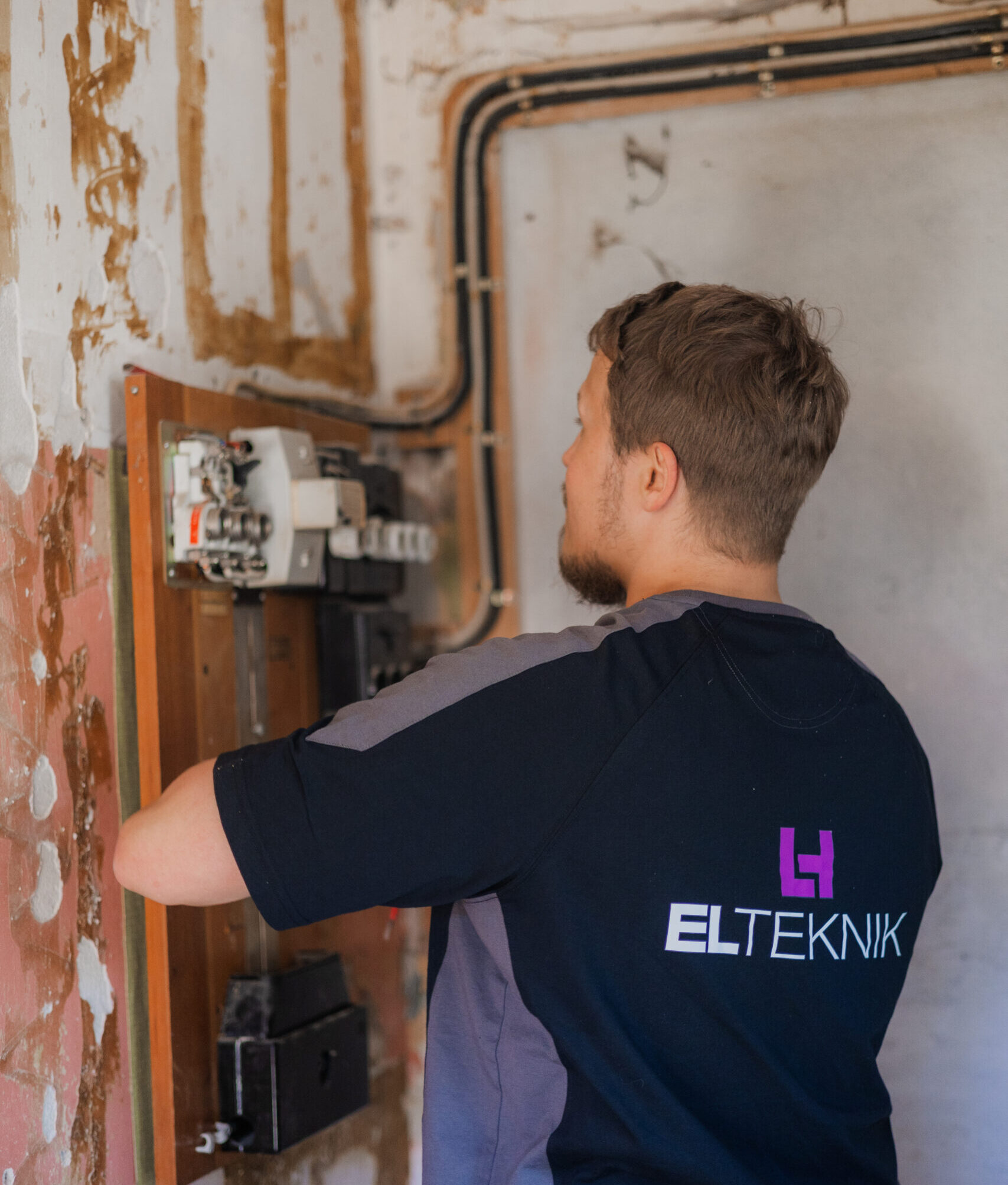 Elektriker fra LH Elteknik arbejder ved en eltavle på en rå væg med synlige kabler og installationer. Hans fokus på komponenterne viser praktisk elarbejde og installation i gang.