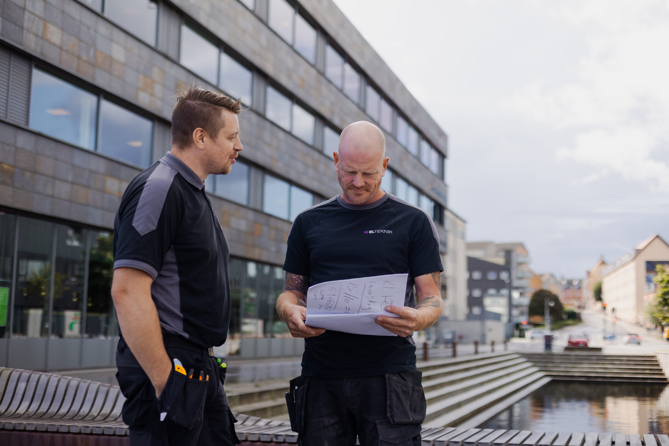 To elektrikere står udendørs ved en moderne bygning og gennemgår tegninger på et papir med skitser og mål. Den ene peger og forklarer mens den anden lytter, hvilket viser planlægning og samarbejde i deres arbejde som elektrikere.