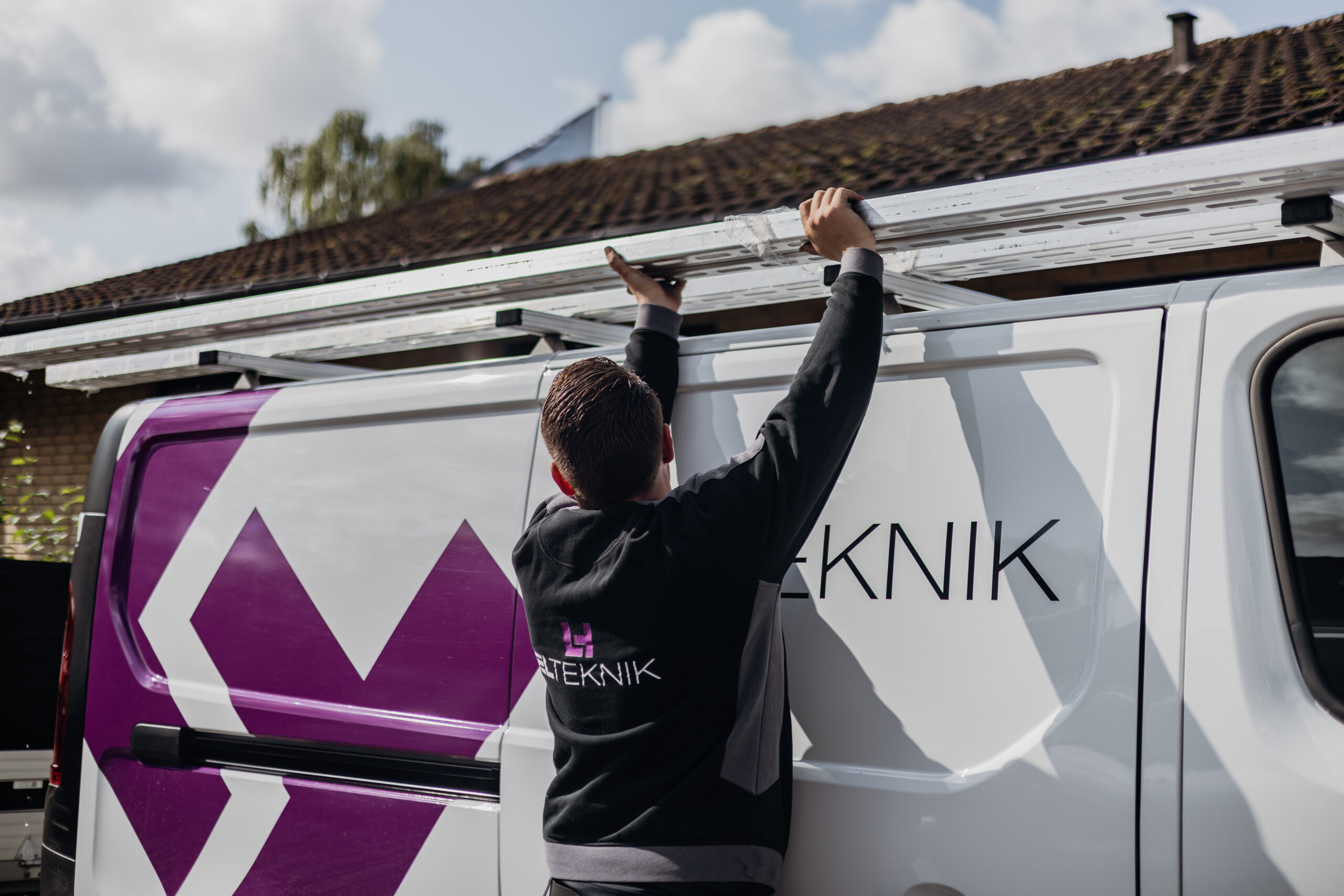 Elektriker fra LH Elteknik løfter en stige op på taget af en hvid servicebil med logoet "ELTEKNIK" på siden. Handlingen viser forberedelse til en opgave som elektriker ude hos kunde.