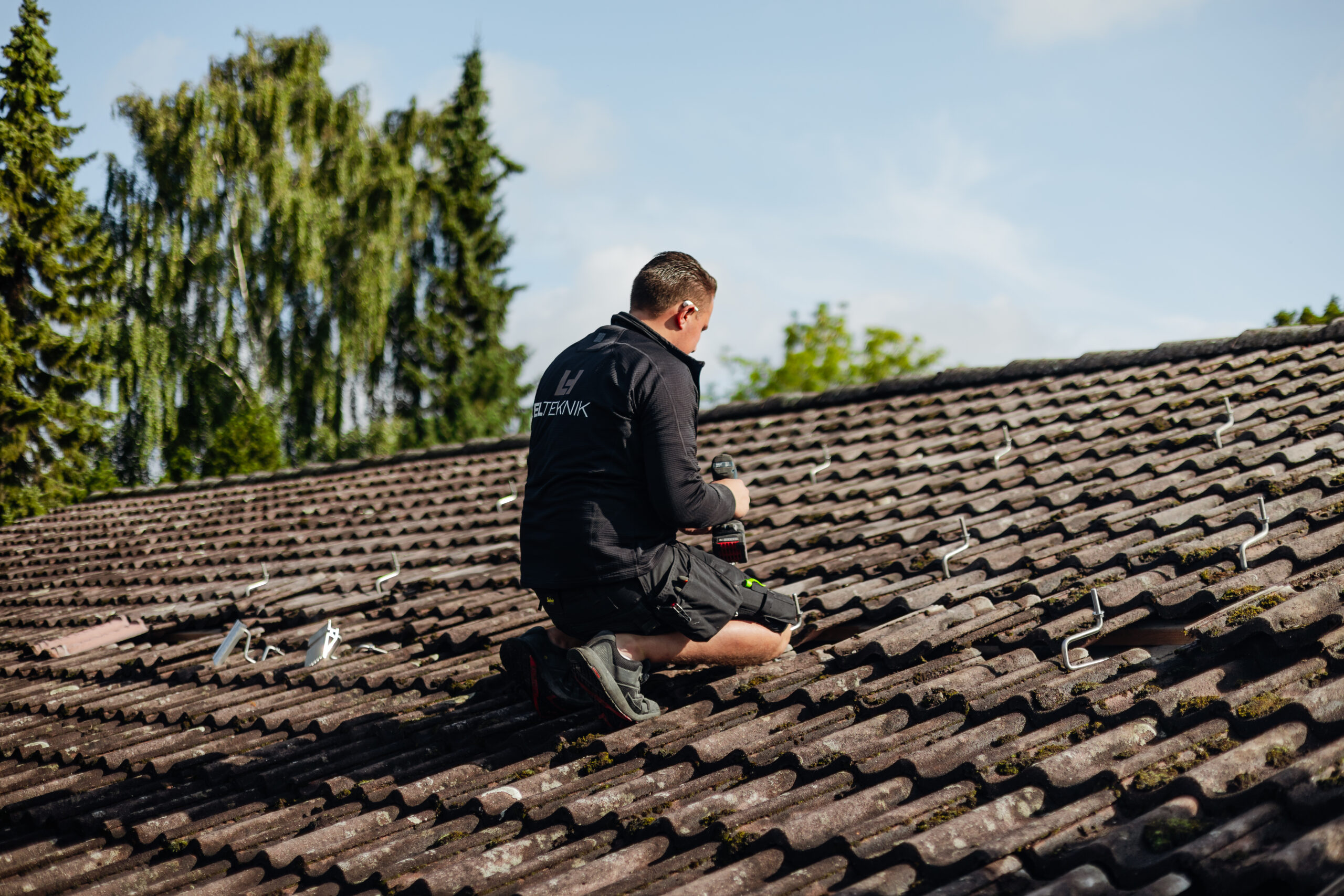 Alt text: Elektriker fra LH Elteknik sidder på et tegltag og monterer beslag med værktøj som led i en installation. De synlige skinner og hans arbejdsposition viser arbejde med elinstallation på tag.