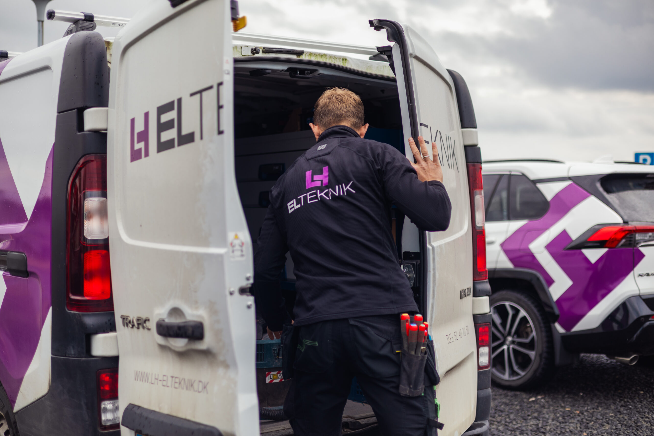 Elektriker fra LH Elteknik står bag sin servicebil med åbne bagdøre og henter værktøj fra bilen før en opgave. Billedet viser forberedelse og organisering af udstyr på arbejdsstedet.