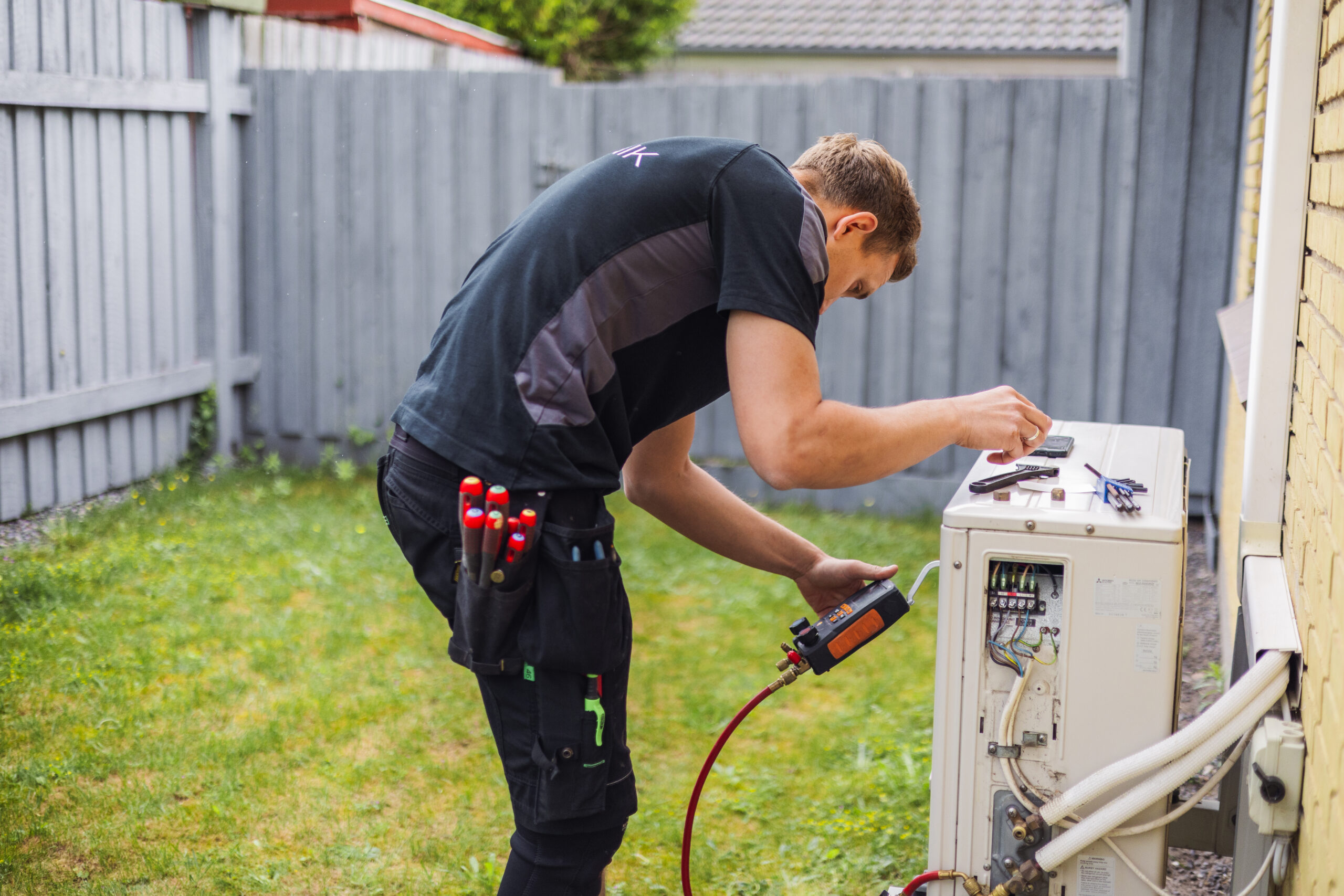 Elektriker fra LH Elteknik arbejder på en varmepumpe udendørs ved siden af et hus og justerer måleudstyr. Billedet viser service og installation af varmepumpe i en privat have.