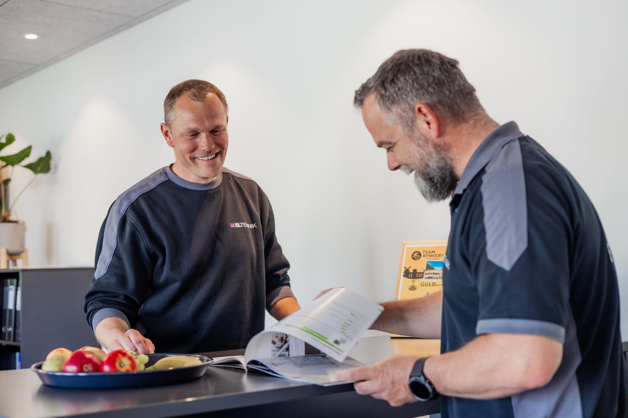 To elektrikere fra LH Elteknik står indendørs ved et bord og gennemgår en brochure sammen med smil, mens de planlægger en løsning. Billedet viser rådgivning og dialog med fokus på kundens behov.