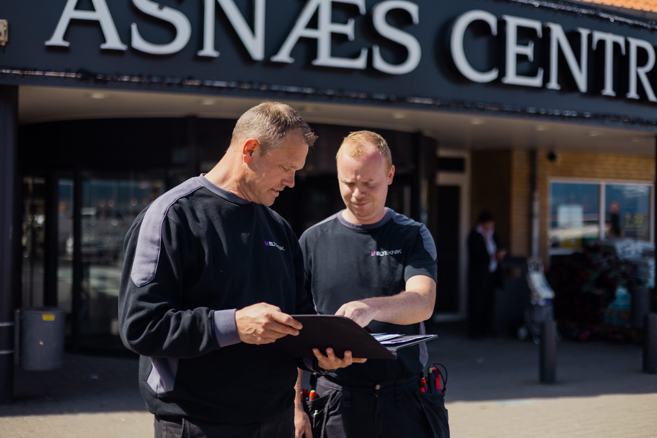 To elektrikere fra LH Elteknik står foran Asnæs Centret og gennemgår en mappe med dokumenter sammen. Billedet viser planlægning af elopgave i bymiljø ved "Asnæs Centret".