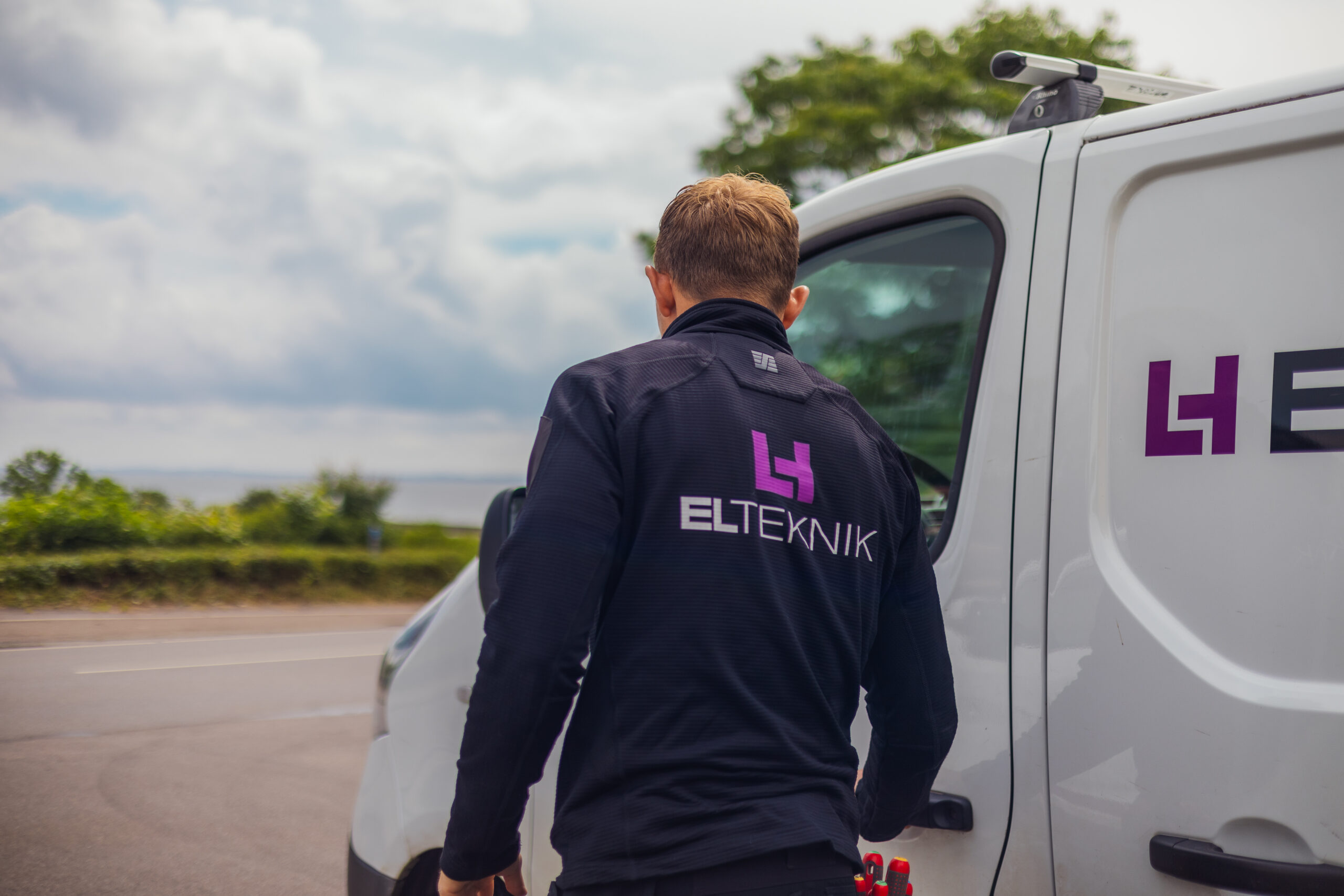 Elektriker fra LH Elteknik går hen mod sin servicebil med ryggen til og udsigt til vand og natur i baggrunden. Billedet viser en tekniker på vej videre til næste opgave i rolige omgivelser.