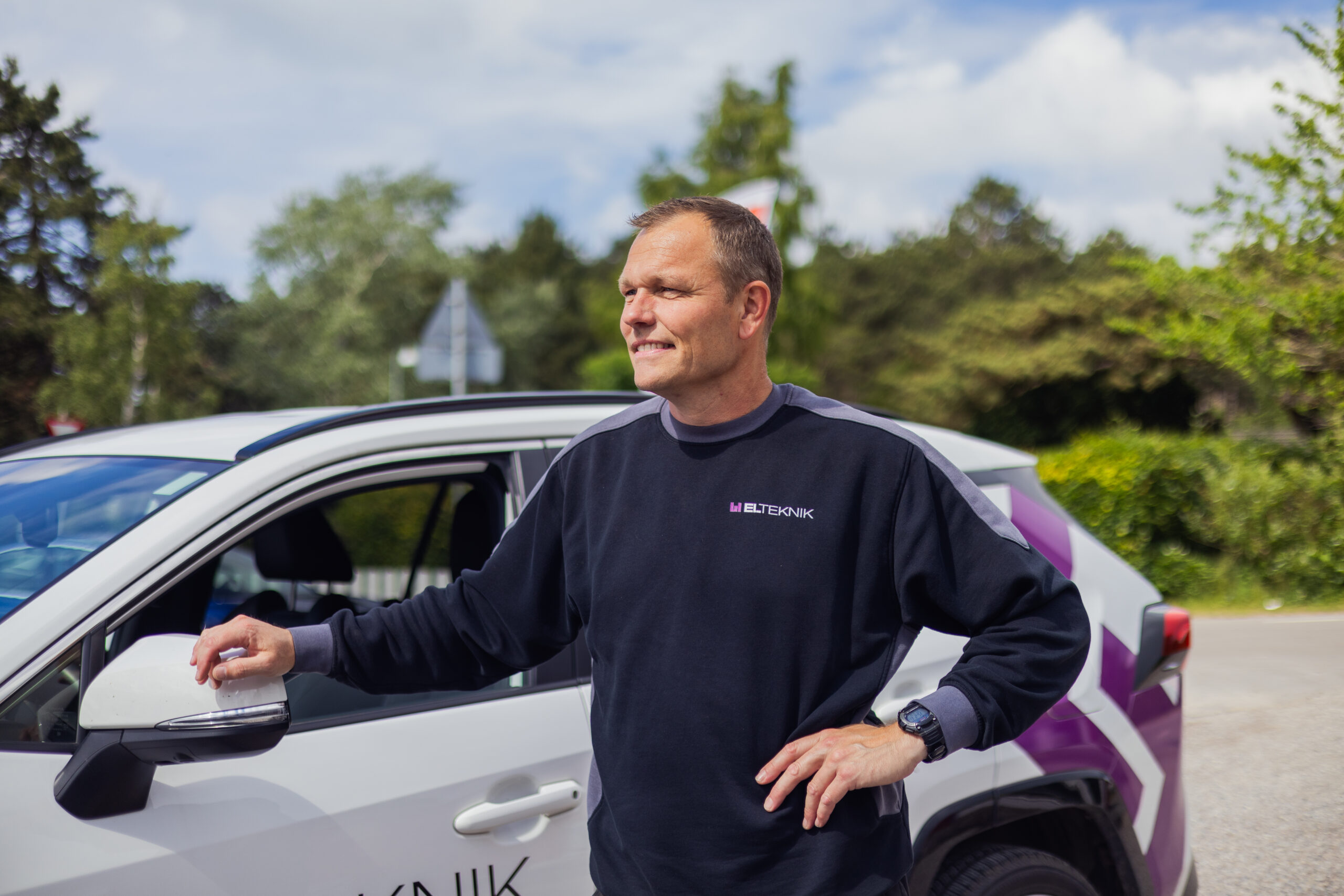 Elektriker fra LH Elteknik står ved siden af firmabilen og kigger ud over området med hånden på sidespejlet. Billedet viser en fagperson klar til opgave ude hos kunde i rolige omgivelser.