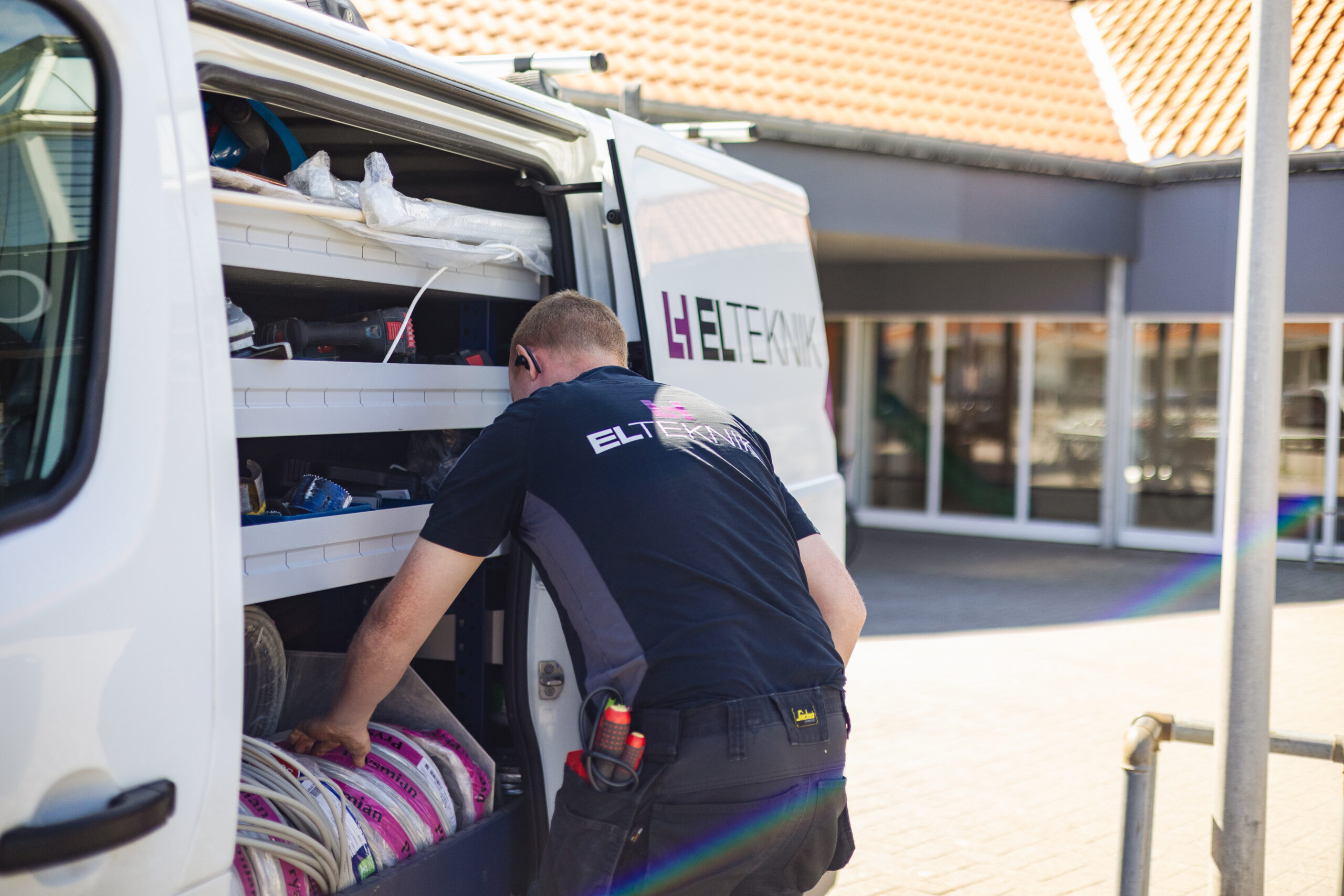 Elektriker fra LH Elteknik står ved sin servicebil og henter materialer og kabler fra de indbyggede hylder bag i bilen. Billedet viser forberedelse til en opgave med organiseret værktøj og udstyr.
