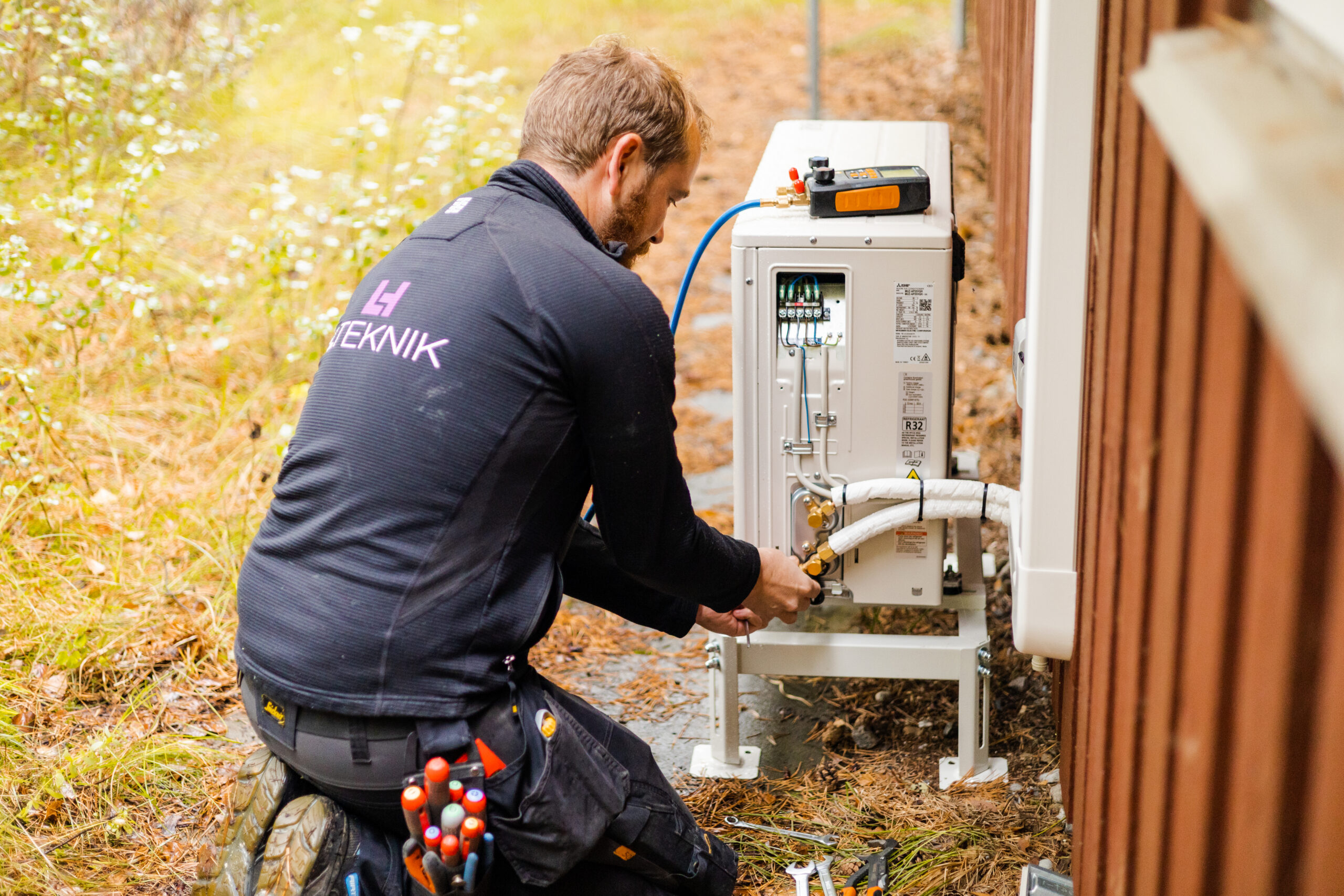 Elektriker fra LH Elteknik arbejder udendørs med installation af en varmepumpe og tilslutter rør og kabler ved enheden ved husets facade. Arbejdet viser opsætning og klargøring af varmepumpesystem til opvarmning.