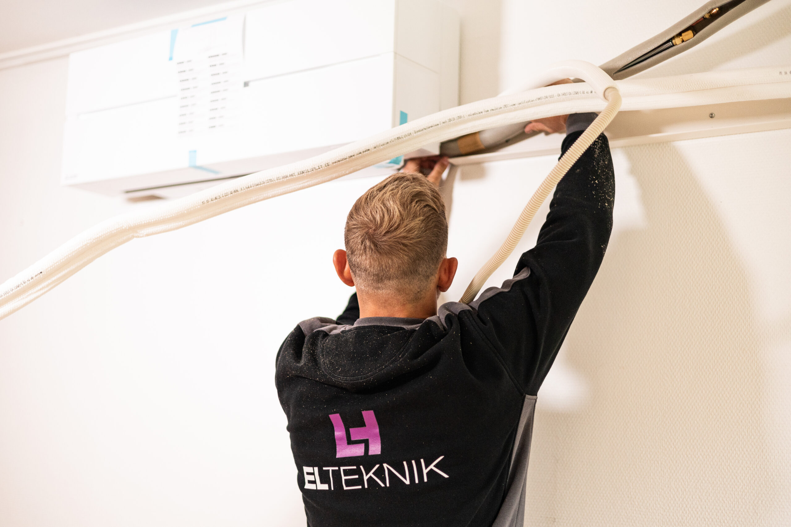 Elektriker fra LH Elteknik monterer en varmepumpe indendørs og tilslutter slanger og installationer langs væggen. Arbejdet viser opsætning af energieffektiv opvarmningsløsning i en bolig.