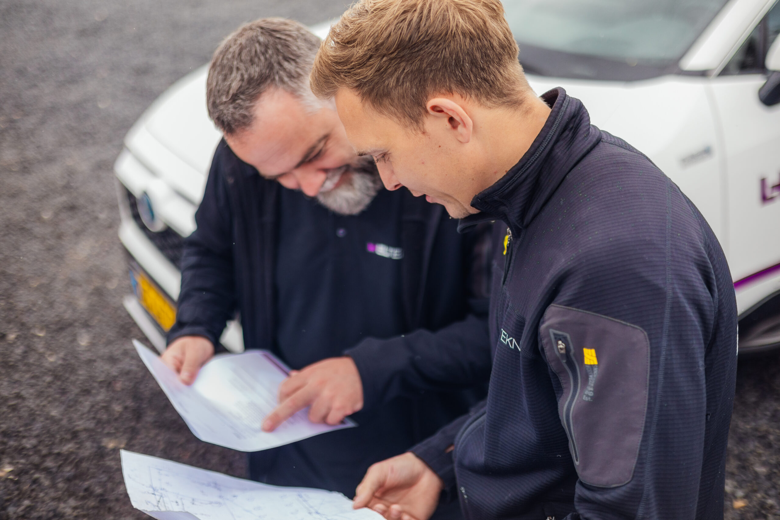 To elektrikere fra LH Elteknik står ved en servicebil og gennemgår tegninger og dokumenter sammen mens den ene peger på papiret. Situationen viser samarbejde og planlægning af en elektrikeropgave på stedet.